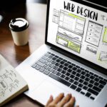 Laptop zeigt Webdesign-Entwürfe, während jemand Notizen macht, um eine Website zu planen, passend zum Thema "Visitenkarte oder Serviceportal".