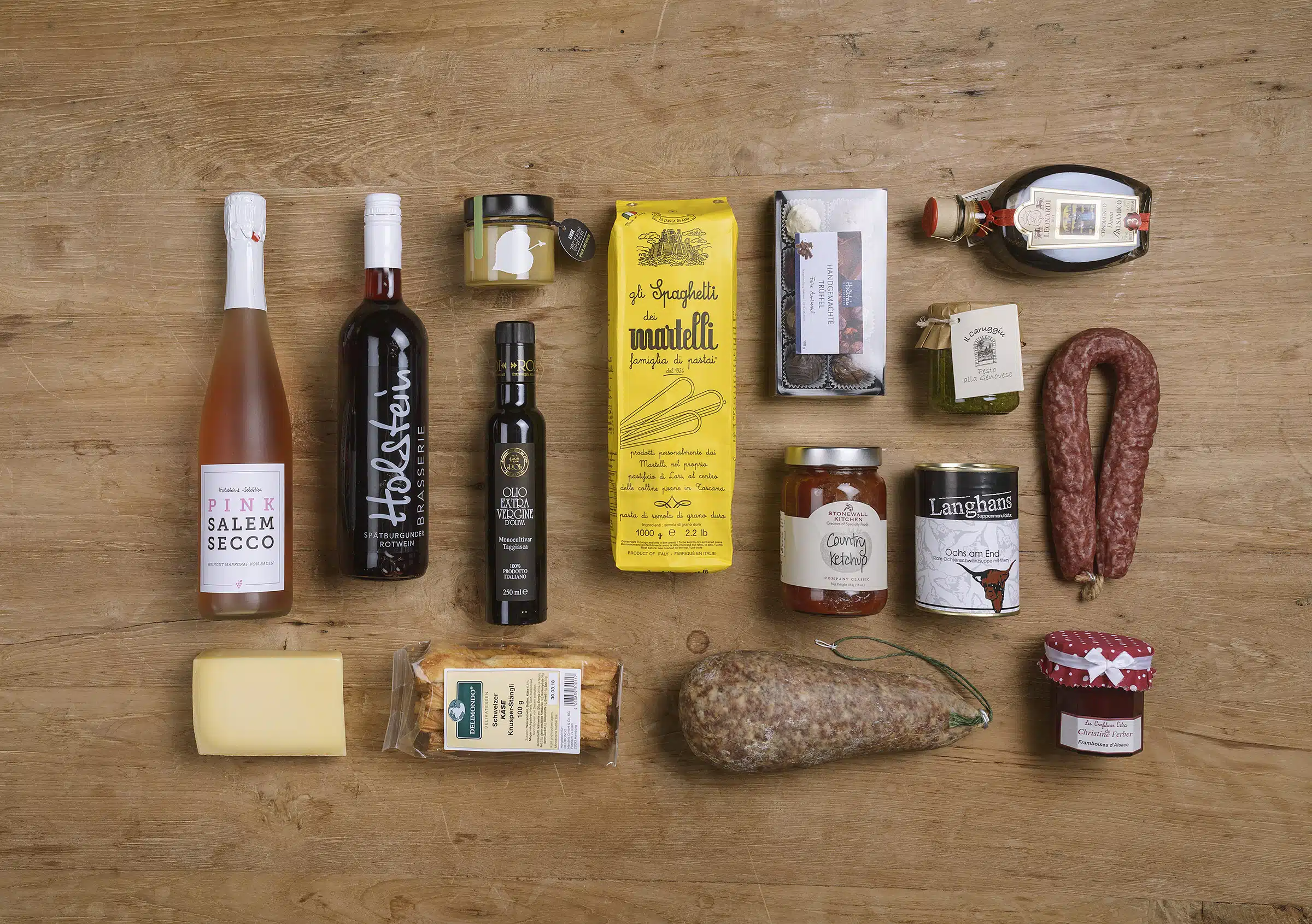 Eine Auswahl von Gourmetprodukten, darunter Wein, Pasta, Olivenöl, Käse und Wurst, auf einem Holztisch arrangiert. Eine Auswahl von Gourmetprodukten, darunter Wein, Pasta, Olivenöl, Käse und Wurst, auf einem Holztisch arrangiert.