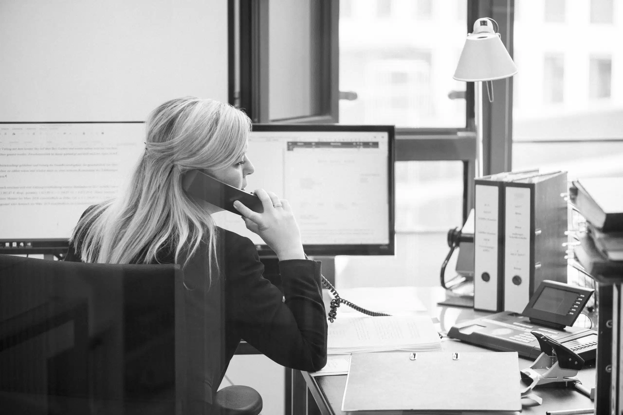 Frau telefoniert im Büro, umgeben von Monitoren und Aktenordnern, mit Blick auf die Stadt durch ein großes Fenster. Frau telefoniert im Büro, umgeben von Monitoren und Aktenordnern, mit Blick auf die Stadt durch ein großes Fenster.